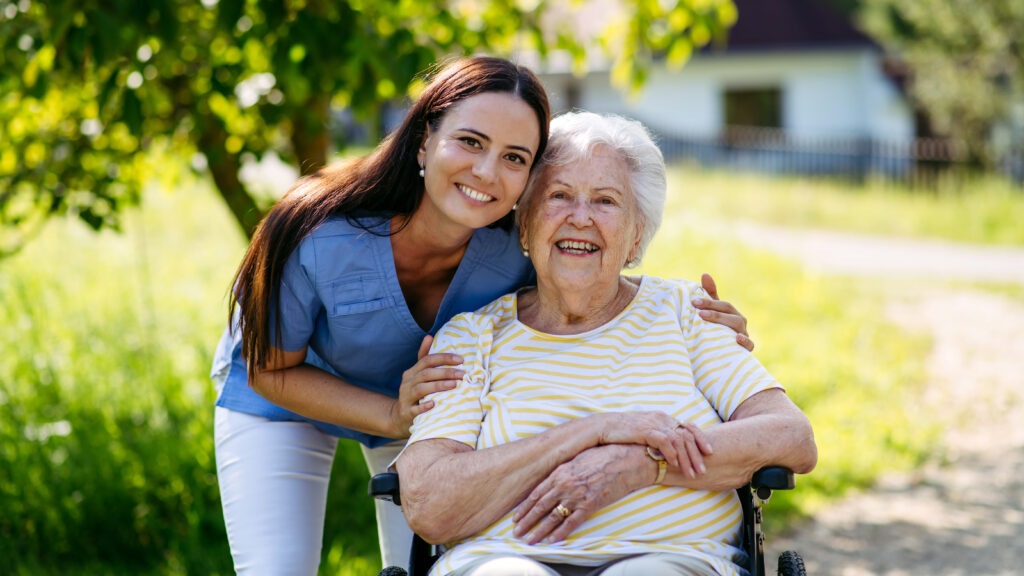 Pflegekraft und Seniorin im Rollstuhl im Gespräch bei einem Spaziergang im Frühling